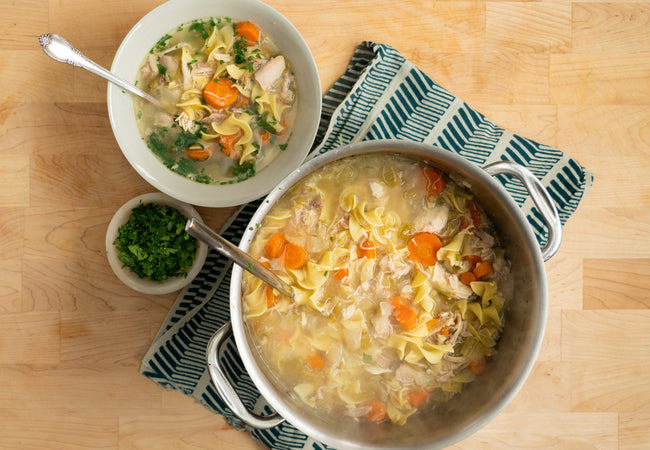 A pot and bowl of pasture-raised chicken noodle soup sitting on a table ready to eat.