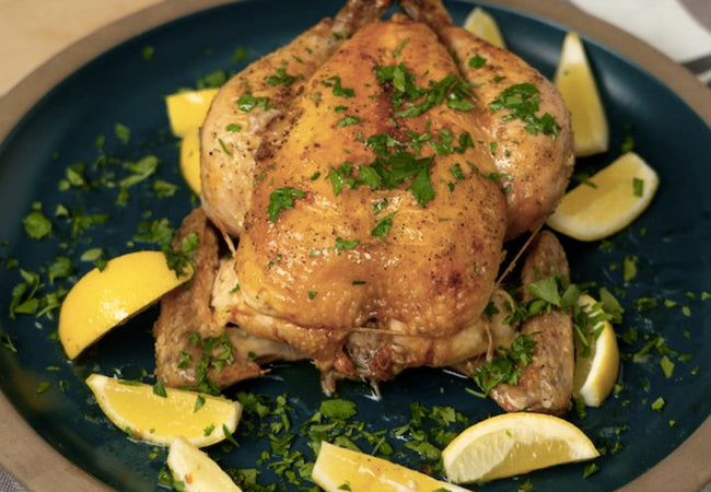 A roasted chicken on a round plate garnish with lemons and herbs.