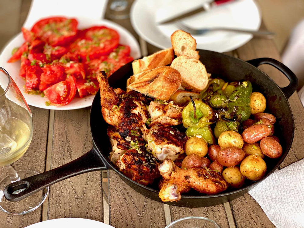 Roasted chicken, potatoes, peppers , and bread in a pan with a side of sliced tomatoes and a glass of wine.