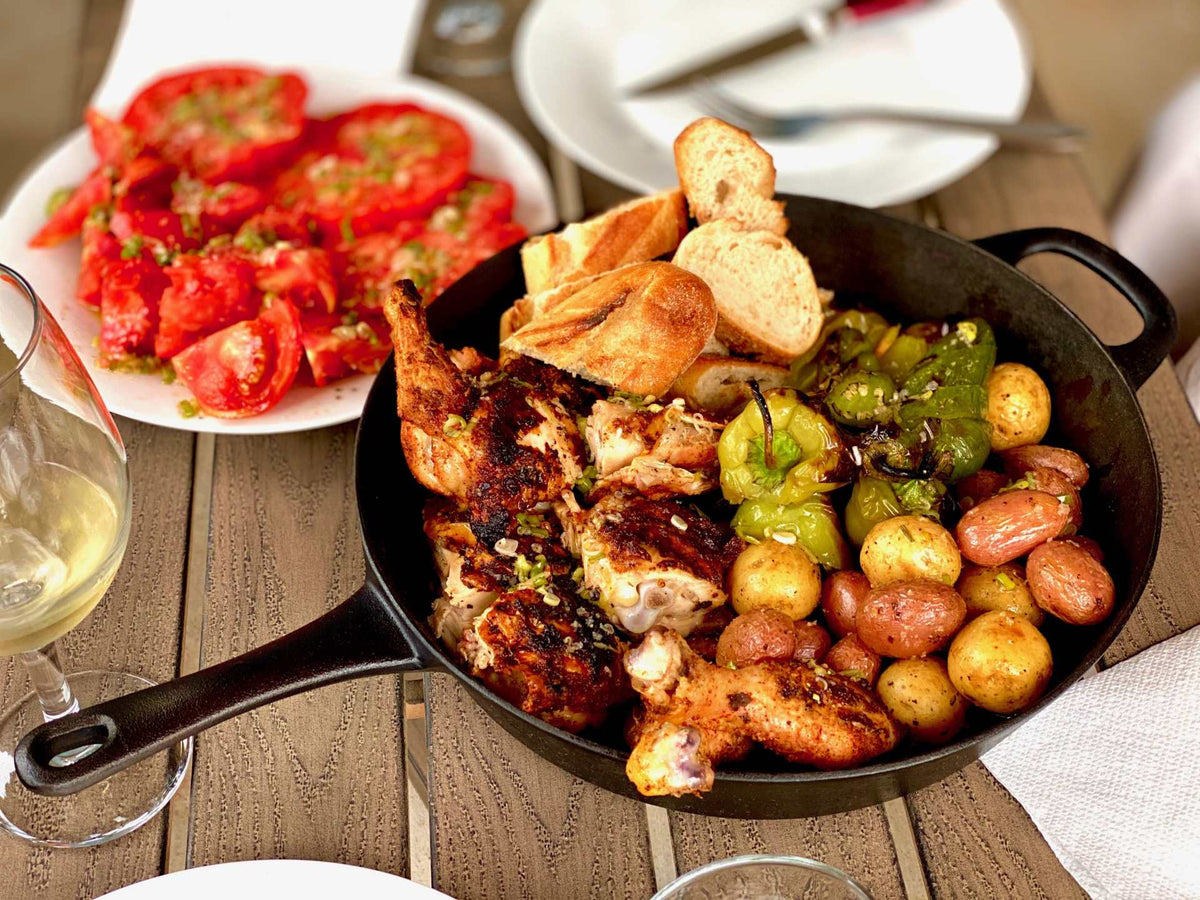 A cast iron skillet with roasted chicken, potatoes, peppers, and bread with a side of sliced tomatoes and a glass of wine.