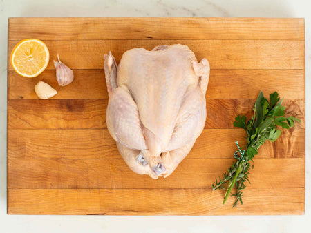Uncooked whole chicken on a cutting board next to lemon and herbs.