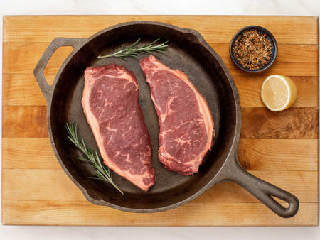 Uncooked New York strip steaks in a cast iron skillet next to seasoning spice and herbs.