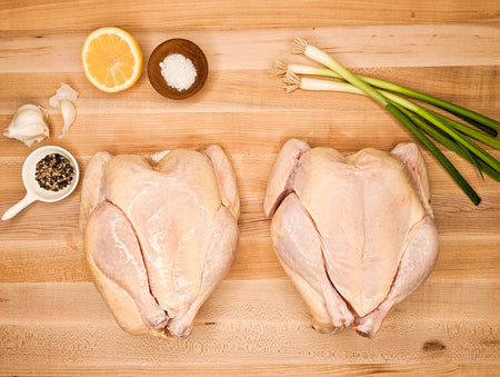 Two uncooked Poussin specialty hens on a cutting board next to seasoning spice and green onions.